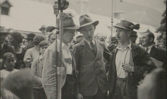 Schwarz-weißes Foto von drei Personen in Tracht an einem Mikro stehend.
