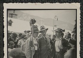 Schwarz-weißes Foto von drei Personen in Tracht an einem Mikro stehend.
