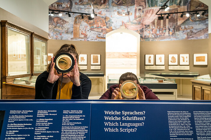 Zwei Personen stehen im Papyrusmuseum und blicken durch eine Lupe direkt in die Kamera.