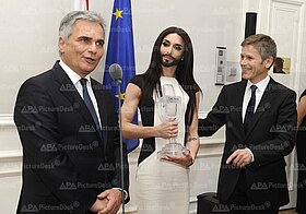 Eine Person mit langen Haaren und Vollbart im Kleid hält den Eurovision-Pokal, rechts und links Personen im Anzug.