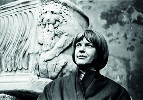 Ingeborg Bachmann vor einem steinernen Löwen, ihr Blick ist nach rechts oben gerichtet.