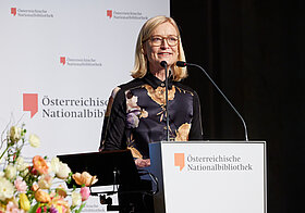 Bebrillte Person am Rednerpult. Im Hintergrund Logowand der Österreichischen Nationalbibliothek.