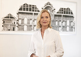 Portraitfoto einer blonden Frau, die vor einem Bild eines antiken Gebäudes steht.