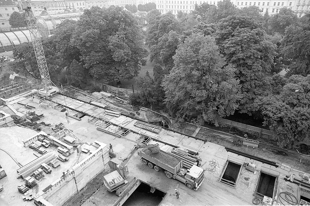 Schwarz-Weiß-Foto einer Baustelle mit einem Traktor, einer Kran und einem gegenüber liegenden Park mit Bäumen.