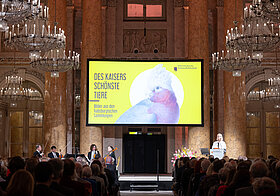 Bühne in einem Marmorsaal mit Lustern, davor Publikum.
