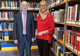 Zwei Personen in eleganter Kleidung posieren vor Bücherregalen mit ukrainischer Flagge.