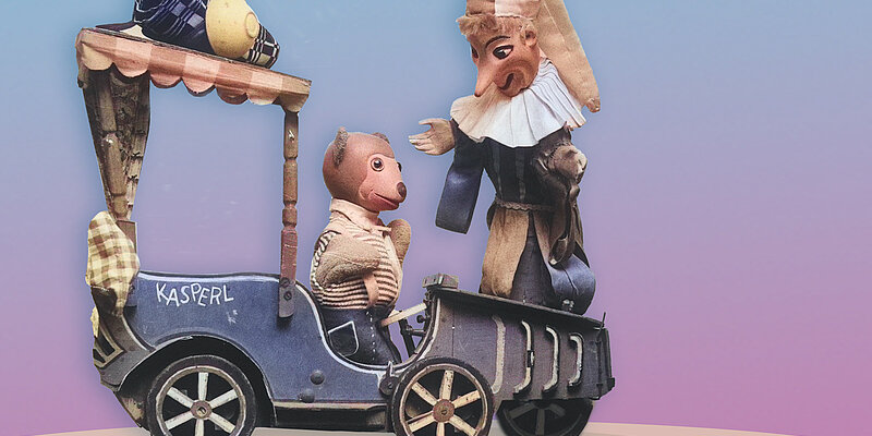 Foto von zwei Handpuppen, eine ist ein Bär und sitzt in einer Kutsche, die andere eine Person mit großer Nase.