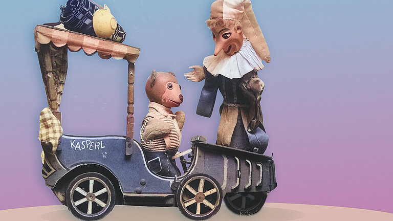 Foto von zwei Handpuppen, eine ist ein Bär und sitzt in einer Kutsche, die andere eine Person mit großer Nase.