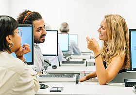 Drei Personen sitzen in einem Computer-Raum und unterhalten sich.