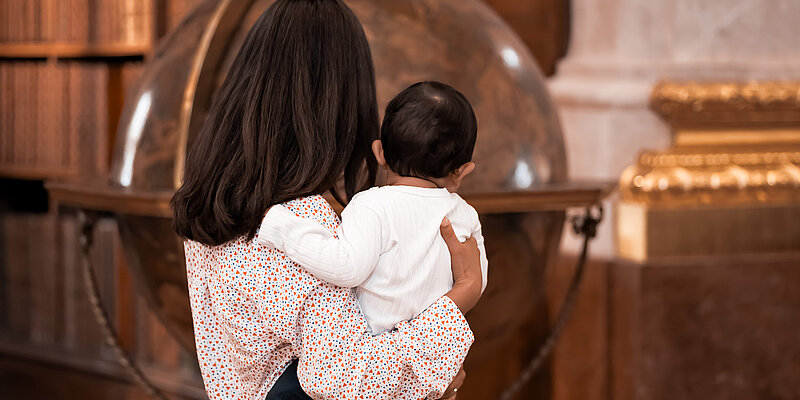 Eine Person mit Baby am Arm von hinten vor einem großen Globus