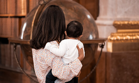 Eine Person mit Baby am Arm von hinten vor einem großen Globus