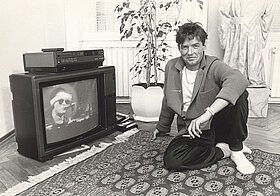 Schwarz-weißes Foto von Person, die in bequemer Kleidung rauchend am Boden auf einem Teppich sitzt und verschmitzt in die Kamera schaut. Daneben steht ein altmodischer Fernseher am Boden, darin wird ein Konzert von derselben Person gezeigt.