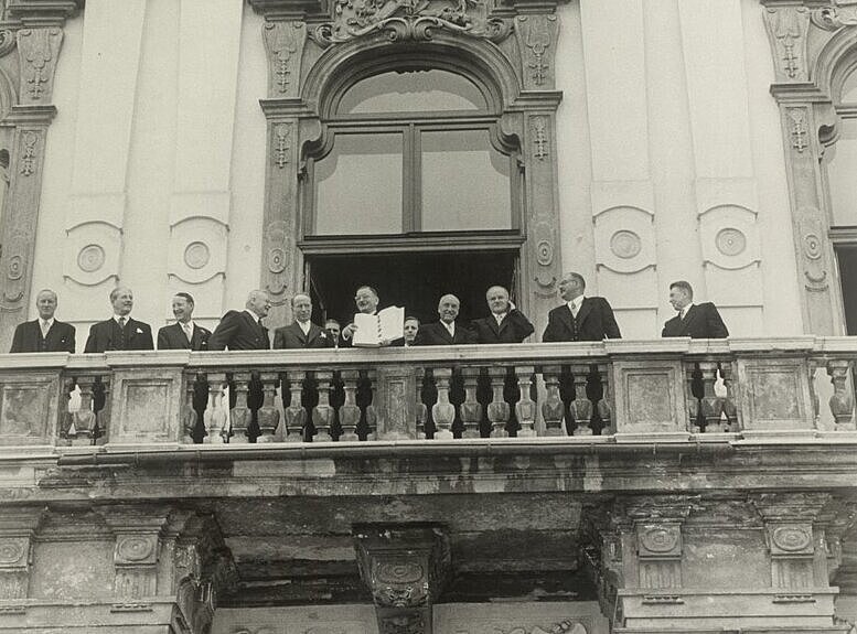Schwarz-Weiß Foto eines alten Gebäudes mit einen Balkon mit vielen Menschen. 