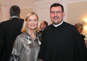 Links eine Person in eleganter Kleidung und eine Person rechts in schwarzer langer Kleidung. 