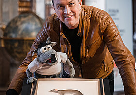 Person und eine Ratten-Handpuppe posieren mit einer Ratten-Aquarellzeichnung.