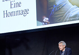Person im Anzug am Rednerpult. Dahinter eine Leinwand mit Ingeborg Bachmanns Foto. 