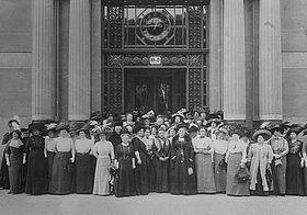 Gruppenfoto von in etwa sechzig Frauen vor einem prunkvollen Gebäude, alle tragen lange Röcke und Hüte, schwarz-weiß.