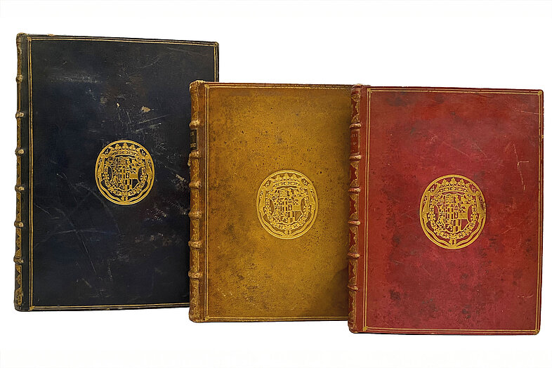 Drei Bücher nebeneinander in Blau, Gelb und Rot mit Wappen darauf.