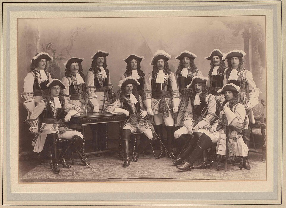 Schwarz-weißes Foto von Personengruppe lockigen Perücken, alle im selben Kostüm mit Piratenhut, weißem Hemd und Degen.