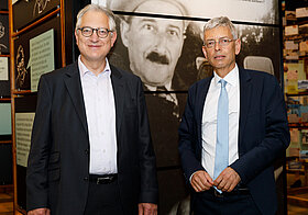 Zwei Personen in eleganter Kleidung stehen im Literaturmuseum. Im Hintergrund ein schwarz-weiß Foto von Stefan Zweig. 