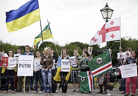 Demonstrierende mit blau-gelben Flaggen und Protestplakaten.