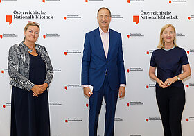 Drei Personen in eleganter Kleidung posieren vor einer Logowand.
