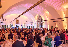 Mehrere Personen sitzen in einem großen Saal auf runden Tischen bei einem Gala-Dinner. 