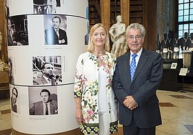  Person mit Blumenblazer und Person im Anzug. Links eine Säule mit schwarz-weiß Fotos.