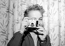 Schwarz-weißes Foto von Person mit kurzen Locken, die lächelnd durch eine Kamera blickt.