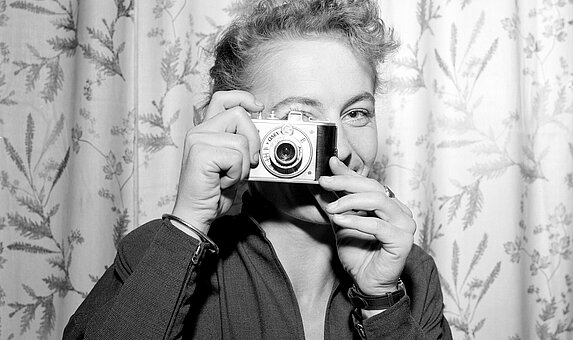 Schwarz-weißes Foto von Person mit kurzen Locken, die lächelnd durch eine Kamera blickt.