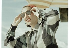 Eine mondäne Urlauberin mit Sonnebrille und Sonneschirm im Hintergrund