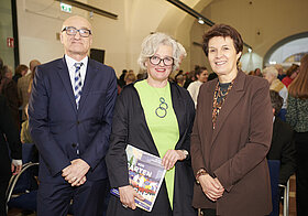 Zwei Personen im Anzug und eine im grünen Kleid hält ein Ausstellungskatalog in der Hand.