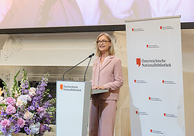 Person in rosa Anzug hält eine Rede an einem Pult, daneben große Blumendekoration.