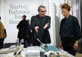 Person zeigt auf eine Schreibmaschine. Eine andere Person steht daneben und schaut aufmerksam zu. 