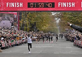 Person läuft durch das Ziel eines Laufs, die Uhr zeigt 1 Stunde, 59 Minuten, 40 Sekunden. Das Publikum jubelt.