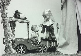 Eine Handpuppe mit langer Zipfelmütze beugt sich über eine kleine Bärenpuppe, die in einem Holzauto sitzt.