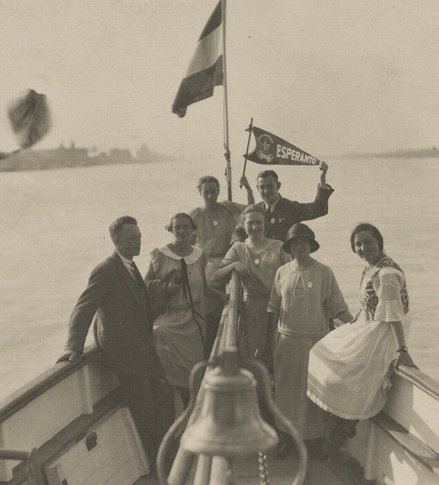 Altes, schwarz-weißes Foto von lächelnden Personen am Bug eines Schiffs am Wasser, eine Person hält eine Esperanto-Flagge.