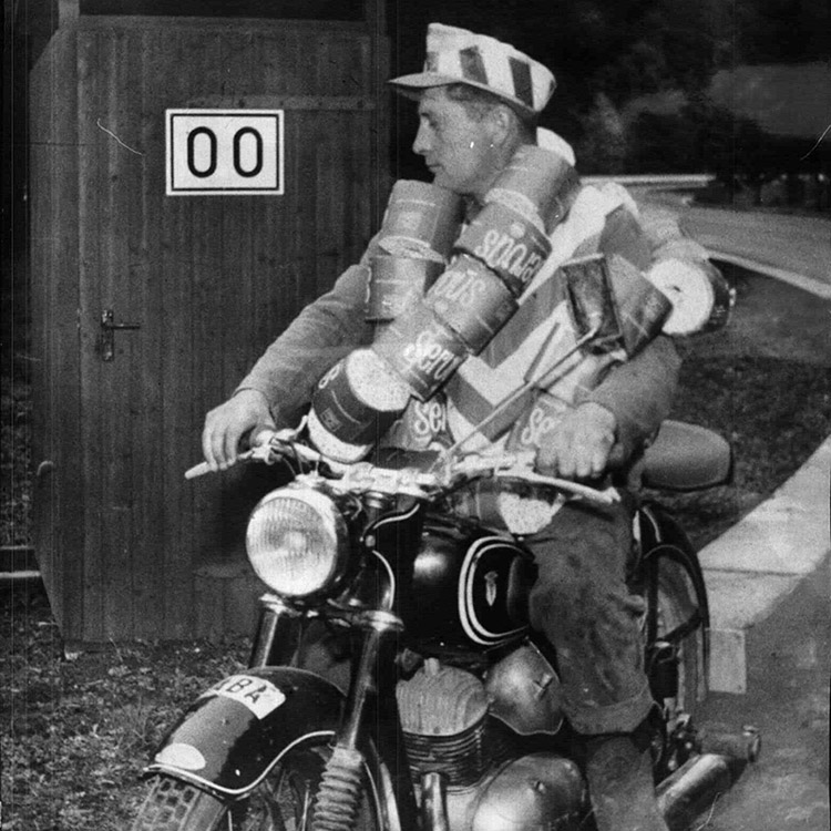 Mann auf einem Motorrad, an ihm sind viele Rollen Klopapier befestigt, hinten steht eine hölzerne Klokabine, schwarz-weiß